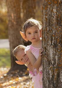 Two fairy girls peering from behind a tree in the Autumn forest