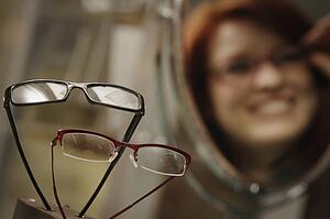 Optician's window display with woman trying on glasses in mirror in background