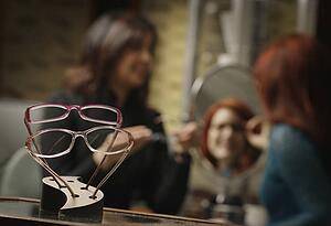 Glasses frames on display with woman trying on glasses with help in the background