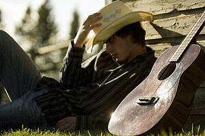 Young man wearing cowboy hat sleeping against exterior of building with guitar