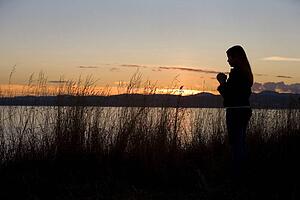 A teenage girl prays at sunset by the ocean.