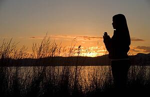 A teenage girl prays at sunset by the ocean.