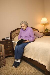 Elderly Caucasian woman in bedroom at retirement community center reading a book.