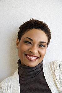 Close up head and shoulder of African- American woman standing against white wall smiling looking at viewer.