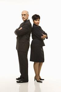 Caucasian middle-aged businessman and Filipino businesswoman standing back to back with arms crossed looking serious.