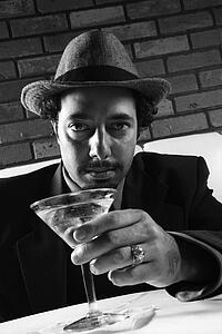 Caucasian prime adult male in retro suit sitting at table with martini looking at viewer.