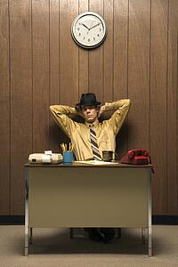 Caucasion mid-adult retro businessman wearing fedora sitting at desk leaning back with hands behind head.