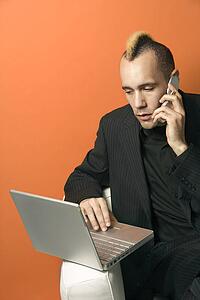 Caucasian man in suit with mohawk typing on laptop and talking on cellphone against orange background.