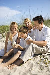 Caucasian family of four sitting on beach looking at seashell.