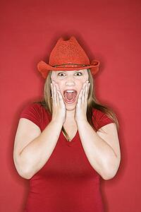 Young Caucasian female adult in red cowboy hat on red background with hands on face and mouth open.