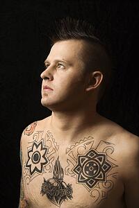 Portrait of shirtless Caucasian man with tattoos and mohawk against black background.