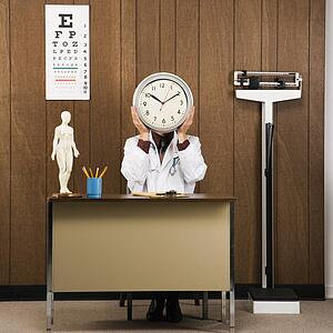 Caucasian male doctor sitting at desk holding clock over face.