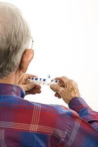 Back over the shoulder view of Caucasion elderly man holding seven-day pill box with Thursday open.