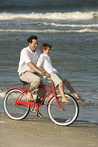 Caucasian mid-adult man riding bicycle on beach with pre-teen boy on handlebars.