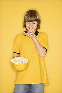 Portrait of Caucasian boy smiling holding bowl of popcorn in one hand and bringing some popcorn to mouth standing against yellow background.