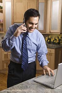 Mid-adult male talking on cellphone and looking at laptop computer in kitchen.