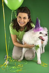 Caucasian prime adult female kneeling with fluffy white dog wearing party hat.