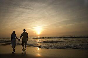 Caucasian mid-adult couple walking holding hands on beach at sunset.
