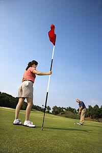 Caucasion mid-adult man putting golfball while Caucasion mid-adult woman holds flag. 