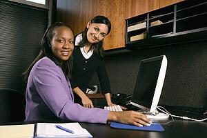 African-American and Indian young adult business women working at computer in office smiling and looking at viewer.