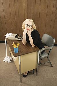 Mid-adult Caucasian female in vintage outfit sitting at desk with chin on her fists.
