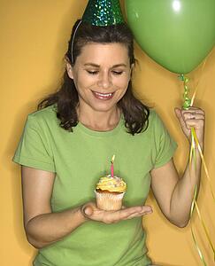 Caucasian prime adult female holding cupcake with lit candle wearing party hat.