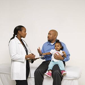 African-American father holding baby girl talking to female pediatrician.
