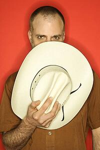 Caucasian man peering over cowboy hat partially covering his face against orange background.