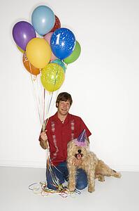 Caucasian young adult male sitting with brown fluffy dog wearing party hat with balloons.