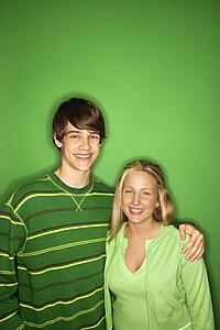 Portrait of Caucasian teen boy and girl standing against green background smiling with arms around eachother. 