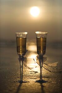 Pair of flute glasses filled with champagne with bows on stems on beach at sunset.