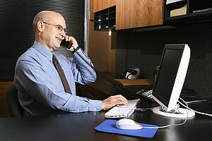 Caucasian middle-aged businessman sitting at desk in office talking on telephone and typing on computer.