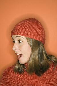Side view of young adult Caucasian woman on orange background wearing winter hat and scarf.