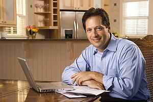 Caucasian mid-adult man paying bills on laptop computer looking at viewer and smiling.