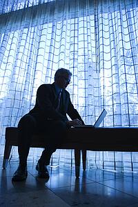Blue tone silhouetted image of prime adult Caucasian man in suit sitting on bench typing on laptop.