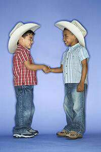 Hispanic and African American male child in cowboy hats shaking hands.