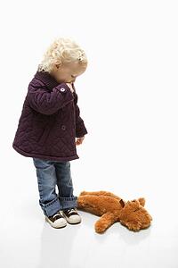 Young Caucasian female toddler sucking her thumb and looking at her teddy bear on ground.