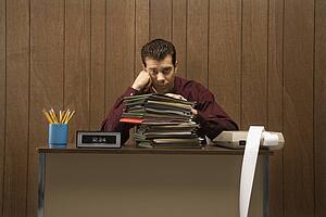 Caucasion mid-adult retro businessman with bored expression sitting at desk piled high with folders.