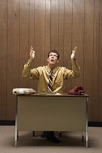 Caucasion mid-adult retro businessman sitting at desk raising hands in air with look of frustration.
