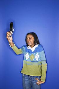 Portrait of African-American teen girl taking photo with camera phone standing in front of blue background.
