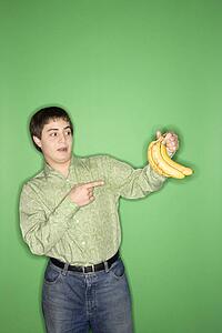 Portrait of Caucasian teen boy holding and pointing at bananas standing against green background.