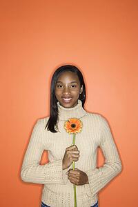 Portrait of African-American teen girl holding single Gerbera Daisy standing against orange background.