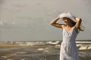 Caucasian pre-teen girl standing on beach holding hat on head.