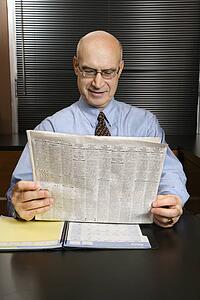 Caucasian middle-aged businessman sitting at desk in office reading newspaper smiling.