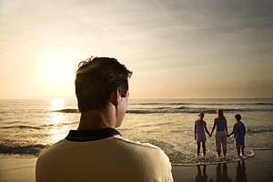 Caucasian mid-adult man standing and watching mid-adult woman with children on beach.