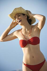 Caucasian mid-adult female in swimsuit holding hat on head laughing.