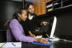 African-American and Indian young adult business women looking and pointing at computer in office.