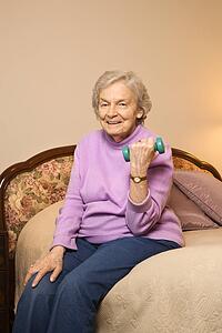 Elderly Caucasian woman in her bedroom at retirement community center lifting weights to strengthen arms.