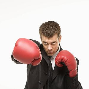 Caucasian mid-adult man wearing suit and punching with boxing gloves toward viewer.