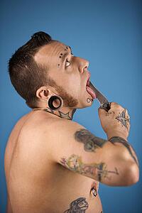 Caucasian mid-adult man with tattoos and piercings holding knife to tongue.